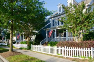 A two-story house with a white picket fence, a front porch, and an American flag hanging by the entrance, surrounded by trees and neatly trimmed shrubs.