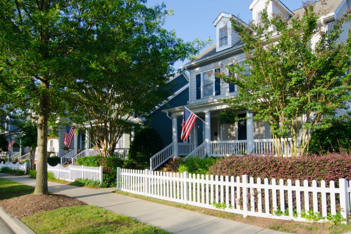 A two-story house with a white picket fence, a front porch, and an American flag hanging by the entrance, surrounded by trees and neatly trimmed shrubs.